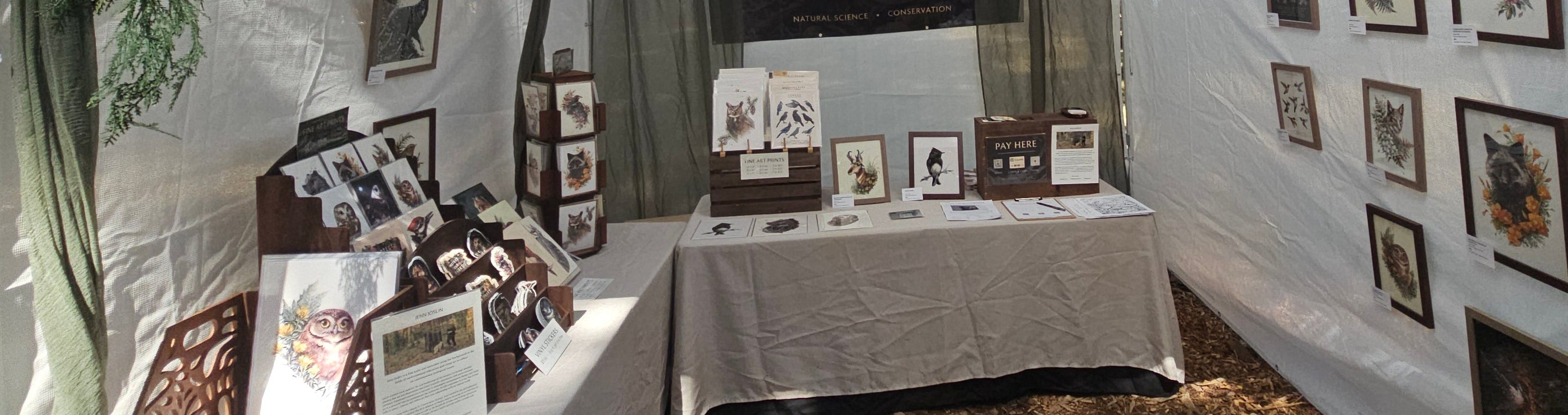 An outdoor market booth under a white canopy tent displaying framed art, photographs, and a table with small items. Trees and a wooded outdoor setting are visible in the background.