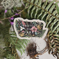 A die-cut sticker featuring a forest illustration with the text 'PROTECT OUR PARKS' and the website 'JENNJOSLIN.COM' at the bottom, placed on a textured fabric surface surrounded by green ferns and dried plant elements.