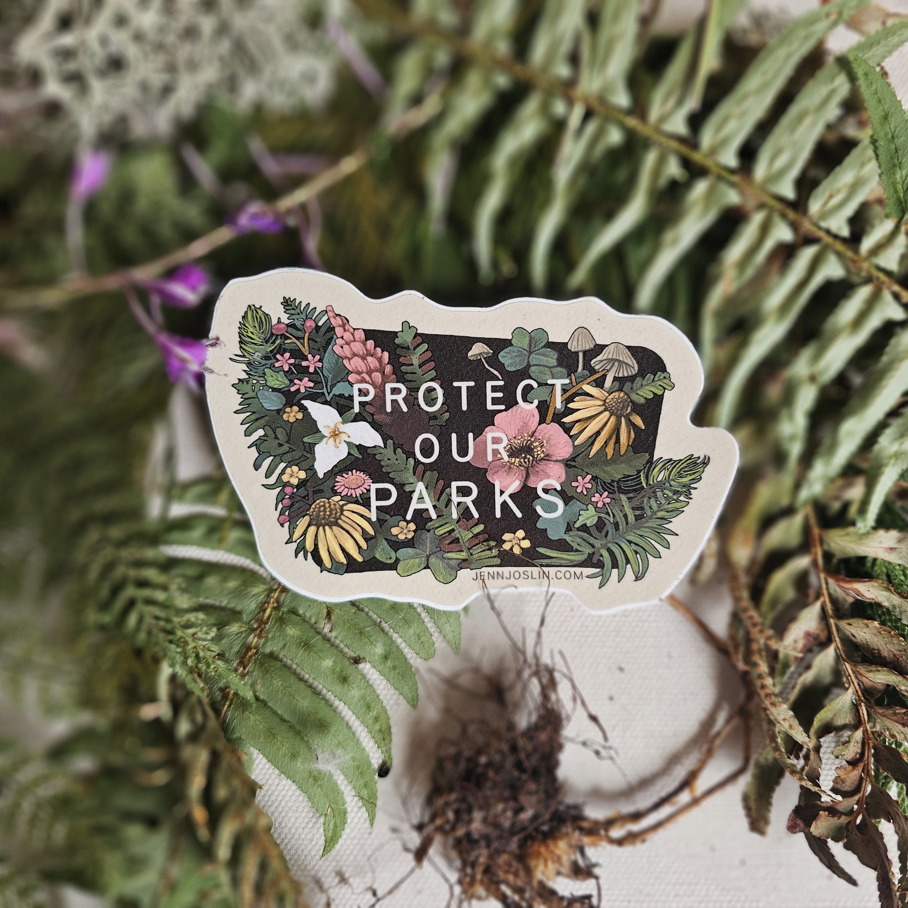 A die-cut sticker featuring a forest illustration with the text 'PROTECT OUR PARKS' and the website 'JENNJOSLIN.COM' at the bottom, placed on a textured fabric surface surrounded by green ferns and dried plant elements.