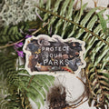 A die-cut sticker featuring an illustration of various animals and the text 'PROTECT OUR PARKS' in white, placed on a textured fabric background surrounded by green ferns and dried plant roots.